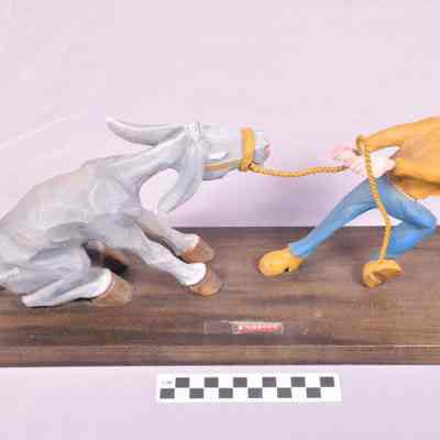 Painted wood carving of mule sitting on haunches being pulled by a man