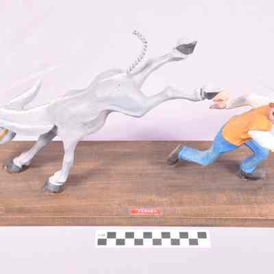 "Perkey" - Painted wood carving of mule with hind legs in air kicking at man who is running away