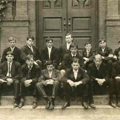 Portrait of fourteen men sitting on steps in front of a building