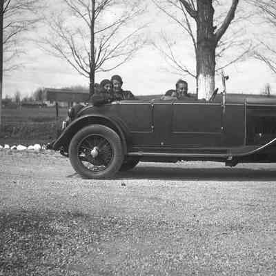 2021.12.310c: Four people sit in a car parked by the roadside, near leafless trees under a clear sky. Dorothy Burnham is in the backseat, behind the driver.