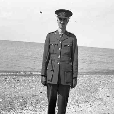 2021.12.310g: A man in a military uniform stands on a pebble beach with the ocean in the background.