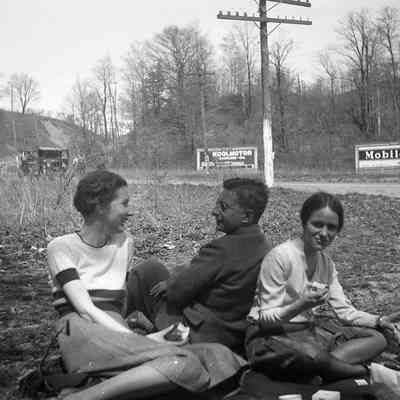 2021.12.310l: Three friends are casually seated outdoors, enjoying a picnic beside a road with Mobil oil advertisements and a vehicle in the background. Printed version of photo is 2021.12.307