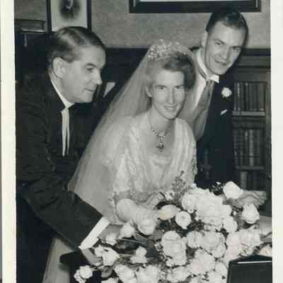 Photo - 2021.12.289c: Dorothy and Harold Burnham pose with a clergyman, surrounded by elegant portraits and florals. Writte on the back, "In the vestry".