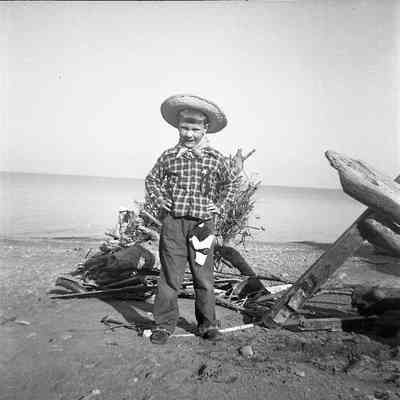 2021.12.309b: A young boy stands smiling on a beach, wearing cowboy attire, a plaid shirt and a straw hat, with driftwood and water behind him.