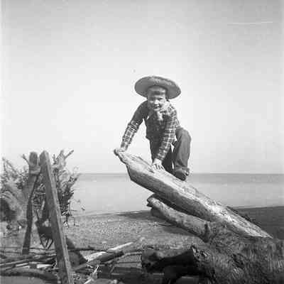 2021.12.309f: A young boy wearing cowboy attire is posed while climbing on a piece of driftwood on a beach.