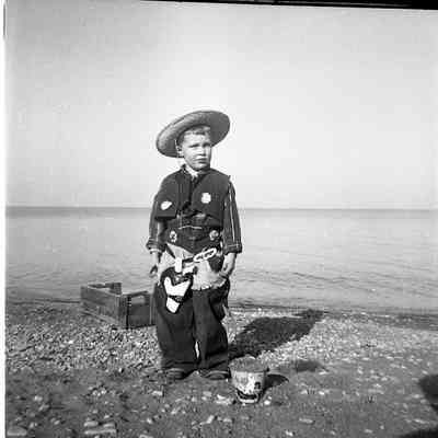 2021.12.309h: A young boy stands smiling on a pebbly beach, dressed in a decorative cowboy costume.