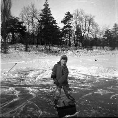 2021.12.309j: A young boy shovels ice on a frozen pond surrounded by trees.