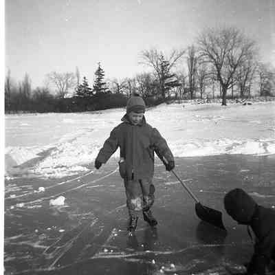 2021.12.309l: A child in a coat skates on a snowy, icy pond, holding a snow shovel.