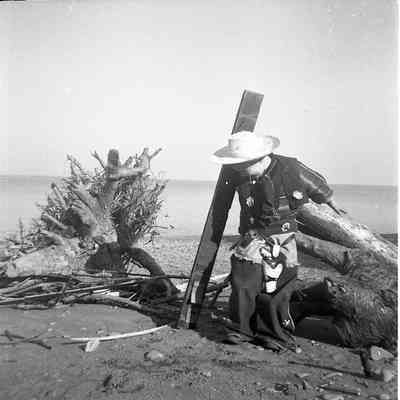 2021.12.309i: A young boy stands in front of a piece of driftwood on a pebbly beach, dressed in a decorative cowboy costume.