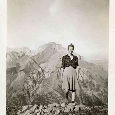 Photograph of Dorothy - 2021.12.291g: Dorothy standing at Sunshine Valley - The Simpson Pass with hills in the background. Photo is attached to black construction paper with two holes punched in it. (Likely part of a scrapbook at one point)