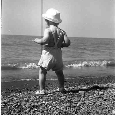 2021.12.311e: A toddler in a white hat stands on a pebbly beach, looking at the sea.