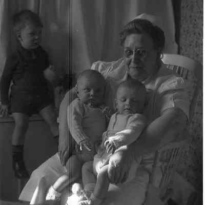 2021.12.311f: A woman sit holding the twins, James and Harold Burnham, while Mark Burnham sits on a window ledge beside them.