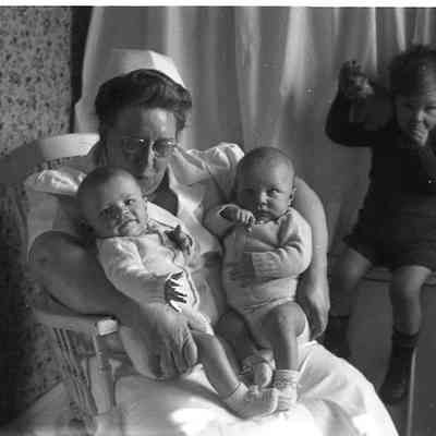 2021.12.311h: A woman sit holding the twins, James and Harold Burnham, while Mark Burnham sits on a window ledge beside them.