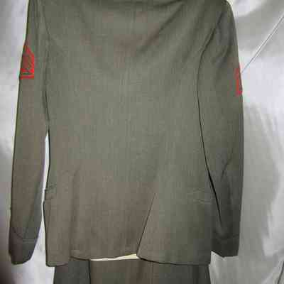 Betty Murray's WWII Marine Uniform back.: The image depicts back side of Betty Murray's WWII Marine Uniform consisting of a dark green buttoned jacket with a beige shirt underneath, decorated with insignia on the shoulders and chest, and a red chevron patch on the sleeve, all hung on a hanger against a plain background.