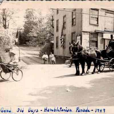 Good Old Days - Hambletonian Parade - 1949: This historical black and white photograph, captioned "Good Old Days - Hambletonian Parade - 1949", captures a closed, horse-drawn carriage driven by a man in formal attire. The carriage, pulled by two horses, rolls down Main Street in front of A.B. Lord's Blacksmith shop, which prominently displays a faded advertisement.