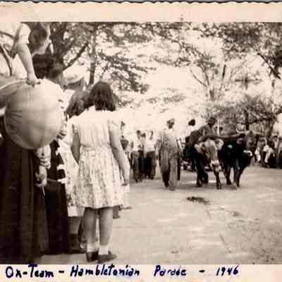 Ox - Team - Hambletonian Parade - 1946 (actually 1949): The black-and-white photograph, dated 1946 (actually 1949), captures a festive moment in the  "Hambletonian Parade," showing Bill Rumsey leading the ox-team on a tree lined street, surrounded by an engaged crowd, including children and someone holding a large, spherical object, possibly a balloon or lantern. The scene evokes a sense of community celebration, with spectators dressed in mid-20th-century attire.