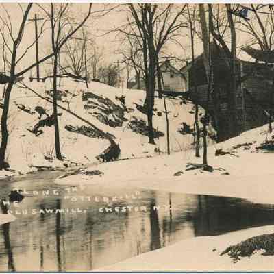 Along the “Otterkill” Old Sawmill, Chester N.Y.: This monochrome photo postcard depicts a serene winter scene along the Otterkill in Chester, New York, featuring a snow-covered landscape with bare trees and a calm, reflective body of water. The right side of the image is bordered by handwritten text, a personal note, reminiscing about these scenes of my childhood. In the background, wooden structures, and buildings of the Old Otterkill  Sawmill, are partially visible through the trees, suggesting an industrial presence amid the natural tranquility.