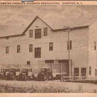 Warehouse of Chester Produce Growers Association, Chester N.Y. Front. chs-019945-3: The image depicts the warehouse of the Chester Produce Growers Association located in Chester, New York, captured in a grayscale vintage style. The sizable two-storied building features a steep, sloped roof and several symmetric windows. In front of the warehouse, a fleet of five antique trucks is parked, each loaded with what appears to be crates of produce, ready for distribution. These vehicles, characterized by their early 20th-century design, hint at the era of the photograph. The rural setting is complemented by a grassy foreground and a partly cloudy sky, which adds to the nostalgic ambiance of the scene. The absence of modern technology emphasizes the historical context of the association’s operations, rooted in a community-based agricultural economy.
