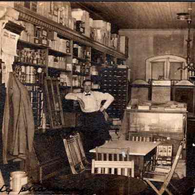 “City Pharmacy Chester NY” Front: The image captures a neatly organized vintage shop or pharmacy from an earlier era, featuring an array of products lined up on high wooden shelves covering the walls. In the center stands a man dressed in typical early 20th-century attire: white shirt, suspenders, and dark trousers, posing with his arms akimbo and leaning slightly against a counter that houses various goods visible through its glass top. Around him, one can see additional furniture such as a table, chairs, and cabinets filled with meticulously arranged items, likely either pharmaceuticals or general store goods. The atmosphere is enhanced by vintage lighting fixtures and the wooden floor, adding to the period feel of the setting. The store appears well-stocked, hinting at the prosperity or thorough maintenance of the business, emphasizing a sense of order and attentiveness to detail.
