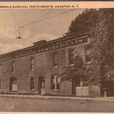 “Plant of Geo. Ermold Surgical Instruments, Chester, N.Y.” Front: The image portrays the exterior of the "Geo. Ermold Surgical Instruments" plant, located in Chester, New York. Featured prominently in the photograph is a sizable, historical brick building, characterized by its arched windows and a distinct signage on the façade that reads "Geo. Ermold Surgical Instruments". Adjacent to the main building are additional structures that seem to be part of the same complex, hinting at their likely use for industrial or manufacturing purposes. A streetlight is visible hanging over the street, subtly indicating the era's urban infrastructure. Furthermore, mature trees partially obscure the right side of the building, adding a touch of nature to the urban scene. This monochromatic photograph captures a moment in time, reflecting the architectural and industrial heritage of early 20th-century America.
