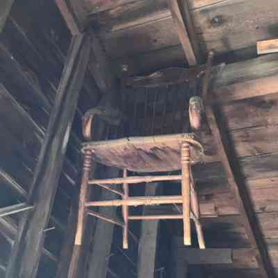 The chairs hung in Ramsdell’s antique store at the corner of Academy Ave and Main Chester NY (aka Durland’s store) for years.: In the image, there is an interior view of a rustic, wooden structure with a slanted roof, where several wooden chairs are precariously stored high above on wooden beams, with one chair noticeably hanging from the ceiling; the walls and ceiling are constructed from weathered wooden planks, some of which exhibit a dark, aged patina, likely reflecting years of exposure to the elements or perhaps smoke from a nearby stove or fireplace, adding to the overall vintage, somewhat eerie atmosphere of the scene; below, there's a clutter of assorted materials, perhaps old doors or panels, stacked haphazardly, suggesting that the space is used as a storage area or attic in an old barn or homestead, offering a glimpse into a forgotten, yet intriguing past frozen in time.