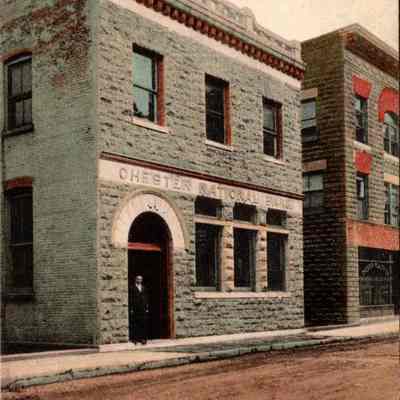 “Chester National Bank, Chester, N.Y.” Front chs-019952-1: The image shows a vintage postcard of the Chester National Bank in Chester, N.Y. This two-story building features a robust stone facade with contrastingly outlined arched entryways and large rectangular windows on the second floor. An ornate cornice tops the building, emphasizing its historical architectural style. A lone figure stands in the doorway, possibly an employee or a patron of the bank. The adjacent building also showcases a similar architectural style but in slightly different colors, adding variety to the streetscape. The color tones of the card are softly muted, typical of early color photographs, set against a sky with dispersed clouds. This postcard captures a quiet moment on a seemingly ordinary day, perhaps in the early 20th century, reflecting the style of that era and providing a glimpse into a historical past preserved on paper.