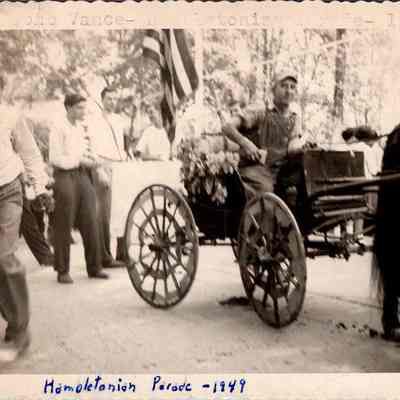 Hambletonian Parade - 1949: The black and white photograph captures the Hambletonian Parade in 1949, featuring a young African American boy in the foreground, walking briskly and looking ahead. In the background, there are several casually dressed spectators, with John Vance driving a flower-adorned horse-drawn carriage.