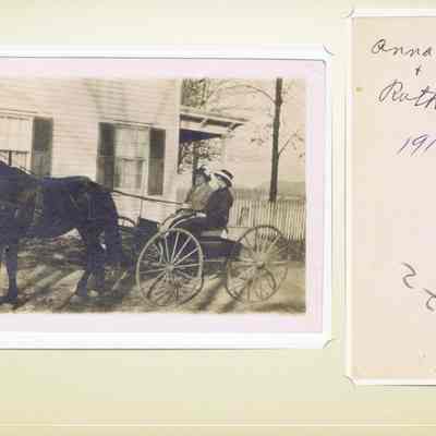 Anna Stedman & Ruth Baird 1916: The image depicts a sepia-toned photograph of a horse-drawn buggy parked in front of a wooden house with shuttered windows, the Baird Home on their farm in Chester NY.  Two individuals sit in the buggy, which is positioned on a dirt path. On the right side of the image, there is a handwritten note on a piece of paper that reads "Anna Stedman & Ruth Baird 1916," alongside the numbers 395 and a small teal-colored strip. The photograph captures a glimpse of early 20th-century life, showcasing the fashion and transportation of that era, with the date and names adding a personal and historical context.