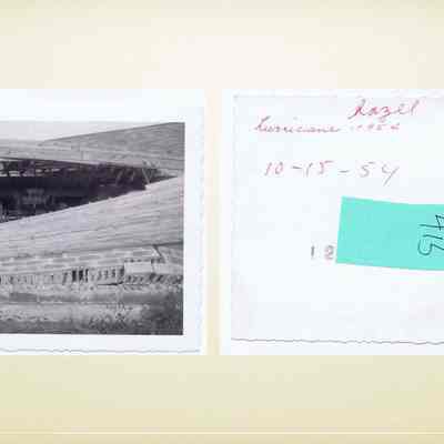Baird Farm after Hurricane Hazel: The image consists of two parts. On the left, a black-and-white photo depicts a barn or shed with a partially collapsed roof, where agricultural machinery, including tractors, is visible beneath the damaged structure. The damage to the building suggests the aftermath of a significant event, likely a natural disaster. On the right side, there is a handwritten note on the back of the photograph, inscribed with "Hazel," "hurricane - 1954," and the date "10-15-54." Additionally, there is a piece of blue paper attached with the number "415" written on it. This suggests that the image documents the destruction caused by Hurricane Hazel in 1954.
