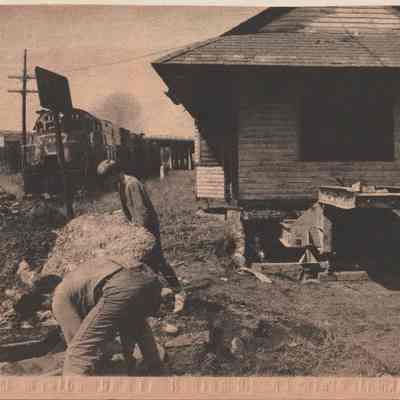 A depot for Sugar Loaf - getting ready to move: The newspaper clipping depicts a scene where a Lehigh and Hudson train is passing by an old site of the East Chester railroad station. Several men from Walter Kannon's moving crew are actively engaged in clearing away debris and dismantling parts of the station, which is still partially intact but being pulled from its foundation. The environment suggests a period of transition, marked by heavy industrial activity and the presence of dated railway equipment.