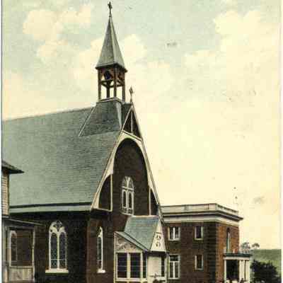 St. Columba's Catholic Church, Chester, N. Y.: This colorized photo postcard depicts St. Columba's Catholic Church in Chester, New York, showcasing a quaint, picturesque example of ecclesiastical architecture from a bygone era. The church, captured in an early 20th-century postcard, features a prominent pointed-arch design with a steep roof and a tall, slender spire adorning the bell tower, typical of the Gothic Revival style. On the right side, an adjacent structure, the associated parochial school with architectural details with children gathered near the school's front entrance.