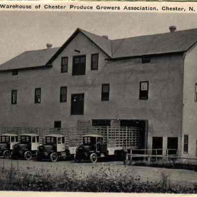20831. Warehouse of Chester Produce Growers Association.: The image showcases the Warehouse of Chester Produce Growers Association located in Chester, New York, featuring a large three-story, stucco-covered building with multiple windows on each level. In front of the building, there are several early 20th-century stake trucks for transporting produce, highlighting the agricultural productivity and transportation methods of that era.