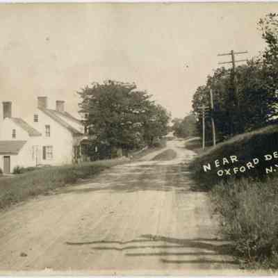Near Oxford Depot, N.Y.: This monochrome photo postcard depicts a serene, early-20th century rural scene near Oxford Depot, N.Y., featuring the Oxford Depot–Craigville Road meandering past, the house of William Henry Woodhull, 1828-1906 with prominent chimneys, lush green trees, and utility poles lined alongside, capturing a quaint and historical countryside landscape.