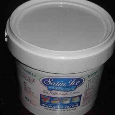 Pastel Green/Vanilla Rolled Fondant 5 lb Container: The image shows a container of Satin Ice Rolled Fondant, prominently labeled as "The Professional's Choice," featuring a secure, snap-on lid in a neutral light gray color; the packaging details its net weight as 5 pounds (2.27 kg), and highlights the item’s intended use in cake decoration and design, showcasing sample images of the product in vibrant, pastel colors on cakes, while providing the website www.satinice.com for further product information.
