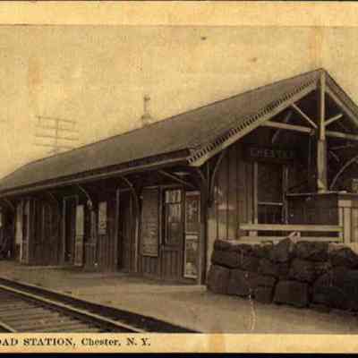 Erie Railroad Station, Chester, N. Y.: This monochrome photo postcard portrays the Erie Railroad Station located in Chester, New York, showcasing a quaint, single-story wooden building with a gabled roof and a large platform, highlighted by rustic architectural details and the absence of any modern elements, evoking a sense of historical transportation practices and simpler times.