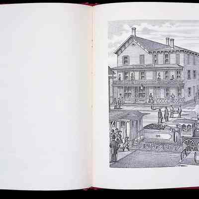 The image displays an open book with a detailed illustration of a bustling 19th century street scene in a chapter titled "Demerest Hall, Warwick." On the right side of the open book, the illustration depicts a large, two-story building labeled "Demerest's Hall" with people and a horse-drawn carriage in front. Adjacent to it is the "National Hotel," also a multi-story building bustling with activity, including individuals on balconies and people entering and exiting. A steam locomotive, labeled with the number 110, moves along the tracks in the foreground, further contributing to the lively street atmosphere of the era portrayed in the book.