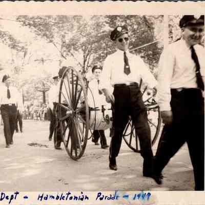 Fire Dept. - Hambletonian Parade - 1949: The black-and-white photograph captures a scene from the " Hambletonian Parade 1949", featuring several men in uniforms, pulling a vintage hand-operated fire hose reel. A woman in period dress watches from the sidelines, while the spectators in the background, including children, add a lively atmosphere to the historical parade event under a canopy of leafy trees.