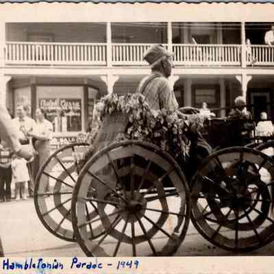 Hambletonian Parade - 1949: The vintage black-and-white photograph depicts a street scene during the "Hambletonian Parade" in 1949,  with John K. Vance wearing a cap and plaid shirt as he drives a flower-decked horse-drawn cart. Onlookers, including a family with children, watch from the sidewalk in front of Ralph Curry's Plumbing and Heating shop while others observe from an upper balcony, creating a lively historic community event atmosphere.