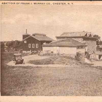Abattoir of Frank J. Murray Co. Chester N.Y. Front: The image presents a historical view of the Abattoir of Frank J. Murray Co., located in Chester, New York. This black and white photograph captures an expansive rural setting with several large buildings that likely functioned as part of the slaughtering and processing operations. The architecture is typical of industrial facilities from the early 20th century, with varying rooflines and what appears to be a mix of stone and wooden construction. In the foreground, a classic automobile adds to the historical ambiance, suggesting the era in which this photo was taken. The landscape is predominantly open fields with scattered vegetation, emphasizing the rural nature of the location. The image serves as a historical document detailing the industrial and agricultural heritage of the region.