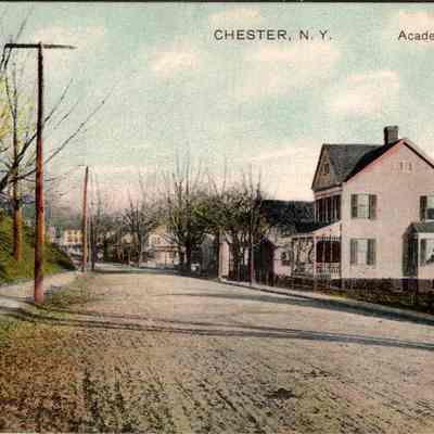 “Chester, N.Y. Academy Ave” Front: This image depicts a nostalgic snapshot of Academy Avenue in Chester, New York, showcasing a serene early 20th-century American streetscape. The road, bordered by pedestrian sidewalks, leads through a residential neighborhood, lined with American-style homes characterized by their pitched roofs, prominent front porches, and varying façade designs. Several bare trees suggest the scene is set in late fall or early spring, enhancing the quaint and quiet ambiance of the town. A gently sloping hillside on the left contrasts with the orderly residential setting on the right. Utility poles stand sentinel alongside the road, reminiscent of a time when such infrastructure was a visible part of everyday life. The image, printed as a colorized postcard, captures both the charm and historical essence of small-town America early in the previous century, inviting contemplation of the lives once lived along this peaceful street.