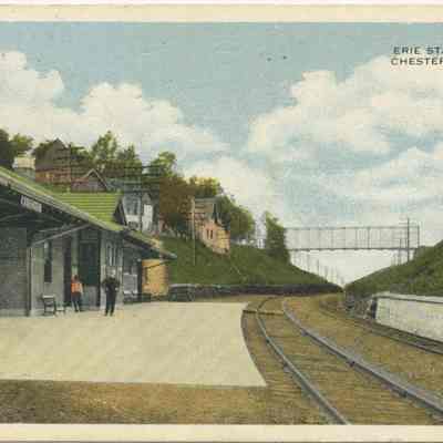 Erie Station, Chester, N. Y.: This colorized photo postcard depicts the historic Erie Station in Chester, New York, featuring a quiet, nearly deserted platform with few passengers. A quaint train station with generous eves to provide shelter, positioned next to a double track leading into the distance under the iron truss bridge spanning the 'Chester Cut" over the Erie RR Mainline Right-of-Way. The serene surroundings include lush greenery, scattered houses on a hillside, and a clear blue sky above, evoking a sense of early 20th-century rural Americana. This postcard captures the nostalgic charm of railway travel from a bygone era.