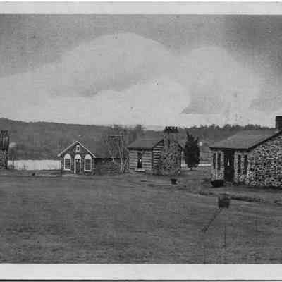 East End of Village Green, Monroe, N.Y.: This monochrome photo postcard depicts a serene landscape featuring a collection of quaint stone buildings scattered across a lush, expansive field, with a backdrop of distant hills partly obscured by a hazy sky, offering a picturesque view that exudes a tranquil, rustic charm at Old Museum Village Of Smith's Clove.