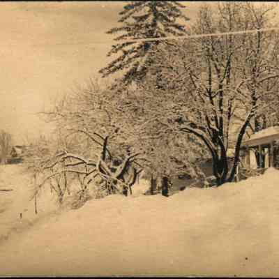 Brook Farmhouse as seen from Main Street: This monochrome photo postcard captures a serene, snowy landscape with houses partially obscured by trees. The Brook Farmhouse on top of the far hill as seen from Main Street.