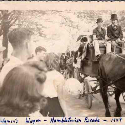 Women's Wagon - Hambletonian Parade - 1949: This vintage black-and-white photograph, captured in 1949, portrays an engaging scene at the Hambletonian Parade, featuring the Women's Wagon. The image captures a crowd of onlookers, some of whose backs are turned toward the camera, watching two men dressed in historical attire, including top hats, seated atop a horse-drawn carriage. One of the men, seated on the left, is Sam Vance.  The carriage and horses are positioned under a canopy of trees, adding a serene, timeless feel to the snapshot.