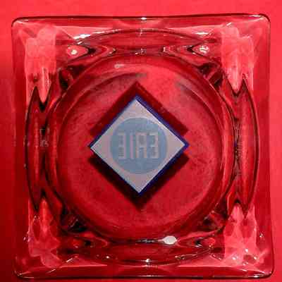 Erie Railroad Square Glass Ashtray - bottom.: Bottom vew of a transparent glass ashtray with a square shape rests on a red background, featuring a blue diamond Erie logo.