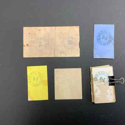 The image displays the back of six rectangular vintage tickets with different colors—faded white, blue, yellow, and tan—each stamped with a circular logo. The logo says "D.L. & W.R.R. CO." with dates from the late 1800s and early 1900s, indicating they are likely historical train tickets. The text within the circles varies, with some saying "Denville," suggesting a location or terminal. The top left ticket, dated July 20, 1899, is quite aged, with visible stains and wear. On the right, a stack of tickets is held together with a black binder clip, suggesting preservation efforts. The arrangement thoughtfully showcases the different designs and aging processes, offering a glimpse into transportation history.