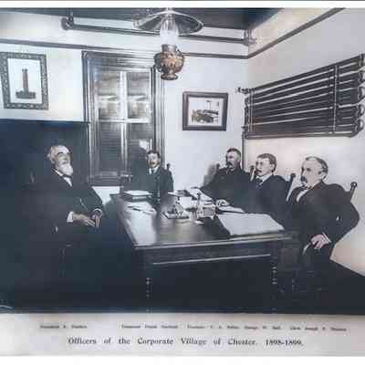Officers of the Corporate Village of Chester 1898-1899. President S. Hadden, Treasurer Frank Durland, Trustees T. A. Miller, George W. Ball, and Clerk Joseph E. Noonan.: The image depicts a historical photograph from the years 1898-1899, showing five men seated around a large wooden table in an office setting, identified as the officers of the Corporate Village of Chester. The room has a high ceiling adorned with a vintage light fixture and features a window with blinds partially drawn. On the walls, there are framed pictures, adding a formal ambiance to the office. The men are dressed in formal attire typical of the late 19th century, including suits and ties, exuding a sense of authority and formality. Each individual has a distinct, thoughtful expression as they engage in what seems to be a meeting or formal discussion. The names and titles of these officers are listed below the photograph: President S. Hadden, Treasurer Frank Durland, Trustees T. A. Miller, George W. Ball, and Clerk Joseph E. Noonan. The atmosphere conveys a sense of historical significance, capturing a moment of governance and civic administration from a bygone era