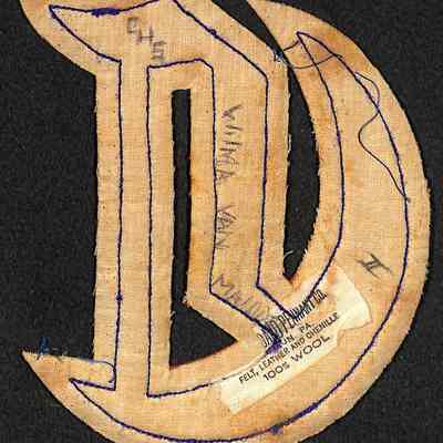Wilma Van Maiden's Chester Letter reverse: The image depicts a fabric patch in the shape of a stylized letter, overlaid with blue stitching, where various handwritten annotations are visible, such as "CHS" and "Wilma Van Maiden," and a manufacturer's label lisitng materials like felt, leather, chenille, and wool, with a percentage noted as 100% wool, all set against a black background which contrasts with the textured fabric and intricate blue thread stitching.
