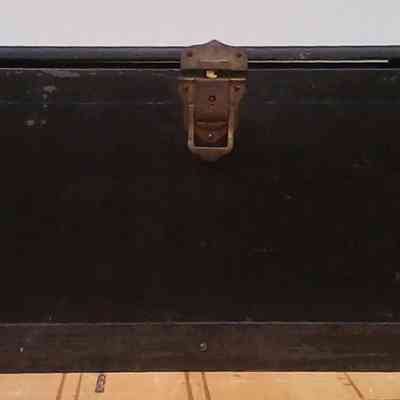 James Razey Box front: A large, old, black wooden trunk with a metal latch rests against a gray wall.