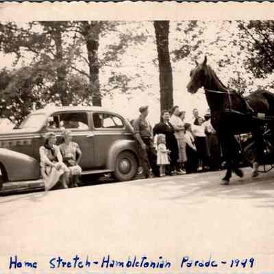 Home Stretch - Hambletonian Parade - 1949: This vintage black-and-white photograph captures a moment from the "Hambletonian Parade", showing a lively street scene where spectators are gathered to watch a horse-drawn carriage. On the left, a group of people, including women in dresses, sitting and standing by an old-fashioned car under the shade of trees, while a man in a suit drives the horse and carriage past the onlookers on the right.